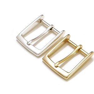May include: Two metal belt buckles, one silver and one gold, are displayed against a white background. The buckles have a rectangular frame with a single prong for securing a belt. The silver buckle is on top, with the gold buckle below.