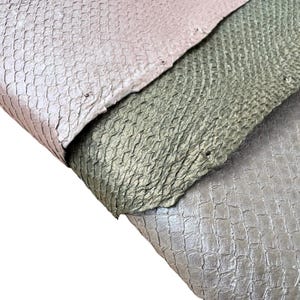 Peut inclure: Gros plan sur trois morceaux de cuir texturé dans les tons rose, vert olive et argent. Le cuir présente un motif en écailles, avec des imperfections visibles et des variations de couleur. Les pièces sont superposées, mettant en valeur les différentes textures.