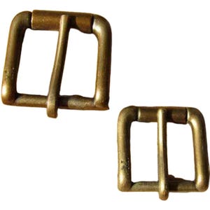 Könnte beinhalten: Zwei antike Messing-Rollschnallen. Jede Schnalle hat eine rechteckige Form mit einem Mittelsteg. Das Metall weist Altersspuren auf, mit dunkleren Bereichen. Einfaches und funktionales Design, geeignet für Gürtel oder Riemen. Aus Messing gefertigt.