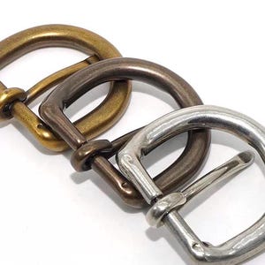 May include: Close-up of three metal belt buckles in brass, bronze, and silver finishes. The buckles are arranged diagonally, showing their D-ring design and varying tones. The background is plain white.