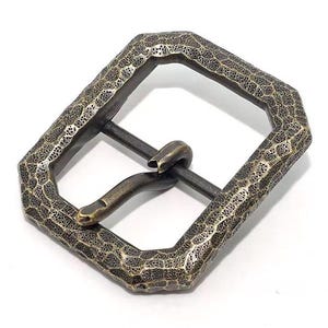 May include: An antique-style belt buckle with a textured, hammered metal finish. The buckle has a geometric shape with a rectangular opening and a hook closure. The color is a mix of bronze and dark grey.