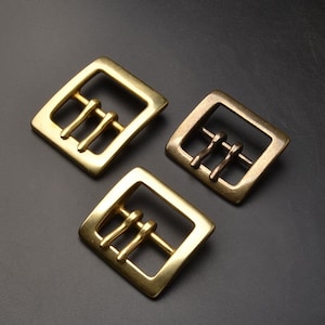 Peut inclure: Trois boucles de ceinture en métal doré avec une forme carrée et trois pointes. Les boucles sont disposées en forme de triangle sur une surface gris foncé.