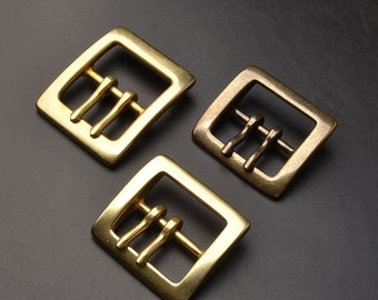 May include: Three gold-toned metal belt buckles with a square shape and three prongs. The buckles are arranged in a triangular pattern on a dark gray surface.