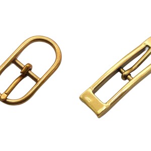 May include: Two gold metal belt buckles. One is oval shaped with a single prong. The other is rectangular with a single prong.
