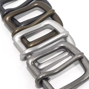 Puede incluir: Una colección de hebillas rectangulares para cinturón en varios acabados metálicos, incluyendo negro, bronce, plata y peltre. Las hebillas están dispuestas en diagonal, mostrando sus diferentes colores y texturas. El diseño es simple y clásico.