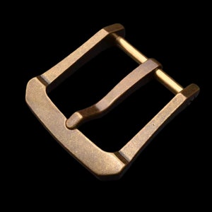 May include: A brass buckle with a smooth, slightly textured finish. The buckle has a rectangular shape with rounded corners and a single, curved prong.