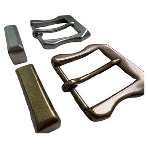Può includere: Primo piano di due fibbie metalliche e due passanti coordinati. Una fibbia è argentata, l'altra è color bronzo. I passanti sono argentati e bronzo. L'hardware è per cinture e altri progetti di pelletteria.
