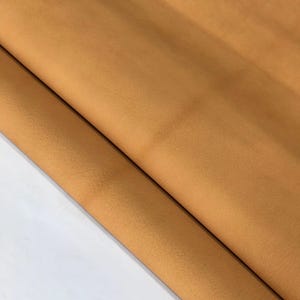 Puede incluir: Primer plano de una hoja de cuero de color marrón claro. El cuero está doblado sobre sí mismo, revelando una superficie texturizada y un tono más claro de marrón claro en la parte inferior. El cuero parece suave y flexible.