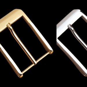 May include: Two metal belt buckles, one gold-toned and one silver-toned, are displayed against a black background. The buckles have a classic rectangular design with a single bar for the belt strap. The gold buckle is on the left, and the silver buckle is on the right.