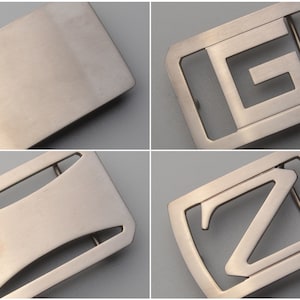 Può includere: Quattro fibbie per cintura in metallo spazzolato. Una è un rettangolo pieno, un'altra ha una "G" stilizzata ritagliata, una terza ha un design curvo e l'ultima ha una "Z" ritagliata. Tutte hanno una forma rettangolare con angoli arrotondati.