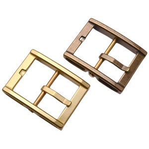 May include: Two metal belt buckles. One is gold-colored, the other is bronze. Both have a rectangular frame with a central bar and a prong for securing a belt. The buckles are shown against a white background.