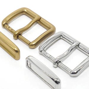 Puede incluir: Dos hebillas de cinturón metálicas, una dorada y otra plateada, con una correa metálica separada. Las hebillas son ambas rectangulares con esquinas redondeadas y tienen un acabado liso.