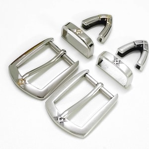 May include: Silver-toned belt buckles and keepers with decorative geometric designs. The buckles have a classic rectangular shape with a single prong. The keepers are curved and designed to hold the belt in place.