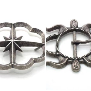 May include: A silver-toned metal belt buckle with a starburst design and a decorative, ornate frame. The buckle has a traditional, functional design with a pin and loop closure.