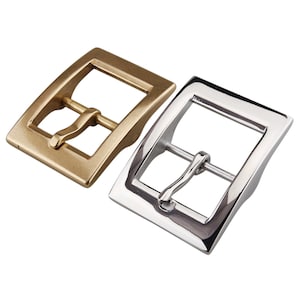 Puede incluir: Dos hebillas de cinturón de metal, una dorada y otra plateada, con forma rectangular y acabado liso.