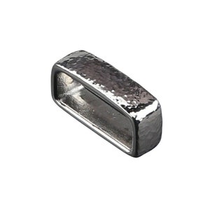 May include: A rectangular, silver-coloured metal buckle with a hammered texture. The buckle has a hollow, open centre and a slightly rounded shape. The metal has a shiny, reflective surface.