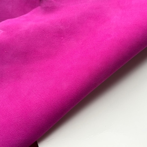 Può includere: Primo piano di un vibrante tessuto magenta con una superficie morbida e strutturata. Il tessuto è piegato, rivelando un effetto a strati e uno scorcio di una tonalità di rosa più chiara. Lo sfondo è bianco.