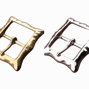 Pode incluir: Duas fivelas de cinto de metal, uma dourada e outra prateada, com acabamento texturizado martelado. As fivelas são quadradas com cantos arredondados.