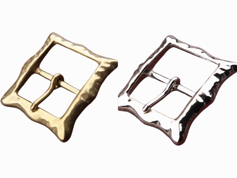 Op de afbeelding: Twee metalen riemgespen, een gouden en een zilveren, met een getextureerde, gehammerde afwerking. De gespen zijn vierkant met afgeronde hoeken.