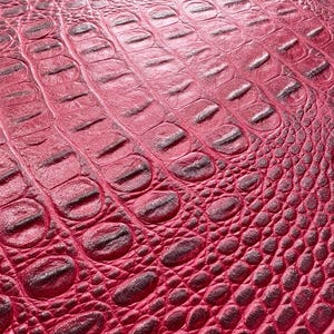 Puede incluir: Primer plano de un material de cuero sintético rojo texturizado con un patrón de piel de cocodrilo o caimán. La superficie tiene un patrón repetitivo de formas ovaladas y rectangulares, con un color rojo más oscuro en las áreas hundidas.