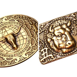 Pode incluir: Duas fivelas de cinto douradas com tema western. Uma fivela apresenta uma cabeça de touro longhorn com o texto "Texas Longhorn Breeders Association". A outra fivela apresenta uma cabeça de touro com intrincados desenhos florais.