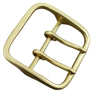 Könnte beinhalten: Eine goldfarbene Metallschnalle mit quadratischer Form und zwei Zinken. Die Schnalle ist für einen Gürtel oder ein Band gedacht.