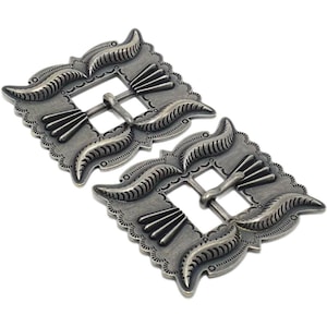 Op de afbeelding: Twee zilverkleurige metalen gespen. Elke gesp heeft een vierkante vorm met decoratieve randen en een centrale staaf. Het ontwerp omvat gestileerde blad- en waaiervormige elementen. De gespen worden tegen een witte achtergrond weergegeven.