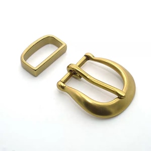 May include: A gold-toned metal buckle with a rectangular keeper. The buckle is designed for a belt or strap.