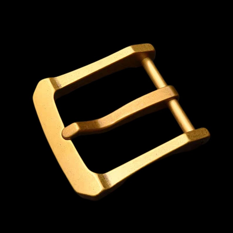 May include: A gold-toned metal buckle with a double-prong design. The buckle is designed for a strap or belt.