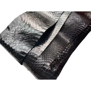 May include: Close-up of several sheets of black, textured leather. The leather has a scale-like pattern and a glossy finish, with visible folds and creases. The material appears to be high-quality and suitable for various crafting projects.