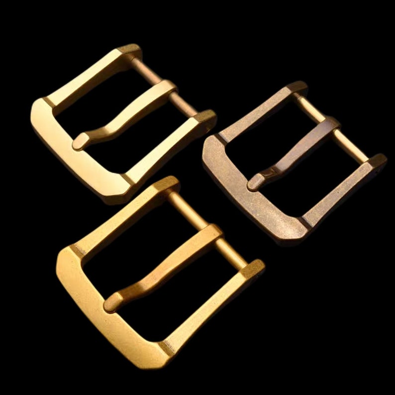 May include: Three metal watch band buckles in different shades of gold. The buckles are all the same design, with a double-prong closure.