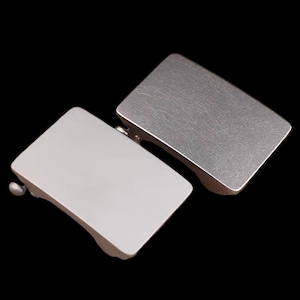 Puede incluir: Dos hebillas de cinturón de metal, una plateada y otra blanca. Ambas hebillas son rectangulares y tienen un acabado cepillado.
