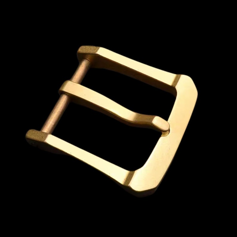 May include: A gold-toned metal buckle with a smooth, polished finish. The buckle has a rectangular shape with rounded edges and a single, centered pin.
