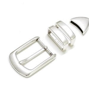 Puede incluir: Un conjunto de componentes de hebilla de cinturón de color plateado, que incluye una hebilla, dos trabillas y una punta de cinturón. Las piezas están dispuestas sobre un fondo blanco, mostrando su diseño y acabado. La hebilla tiene forma rectangular.