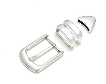 May include: A set of silver-colored belt hardware components, including a buckle, two belt loops, and a belt tip. The pieces are arranged on a white background, showcasing their design and finish. The buckle has a rectangular shape.