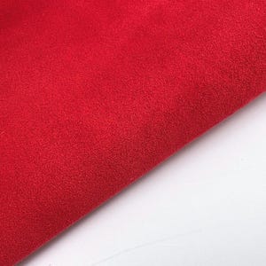 May include: Close-up of a vibrant red fabric with a soft, textured surface. The fabric is folded, revealing a diagonal edge against a white background. The material appears to be a plush textile, possibly velvet or felt.