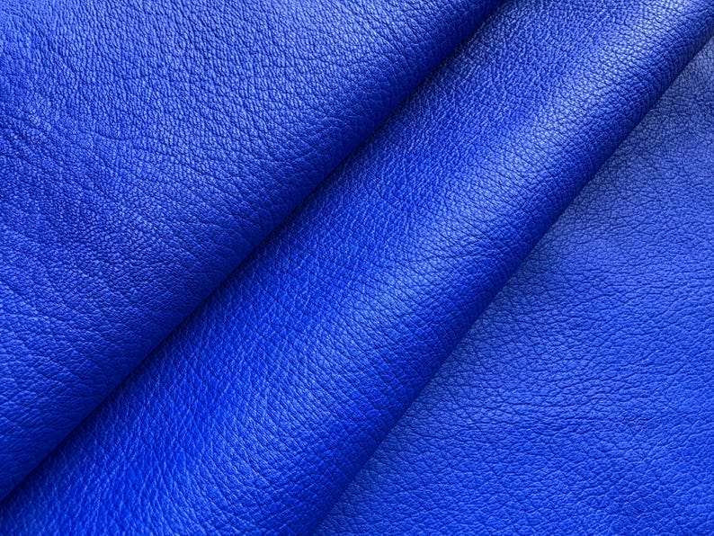 May include: Close-up of vibrant blue leather fabric. The material has a textured surface with subtle variations in color and tone. The folds and creases add depth and dimension to the textile, suggesting its flexibility and softness.