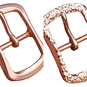 Könnte beinhalten: Zwei roségoldene Gürtelschnallen. Eine hat eine glatte Oberfläche, die andere eine gehämmert Textur. Beide haben eine rechteckige Form mit einem Mittelsteg für den Gürtelriemen.