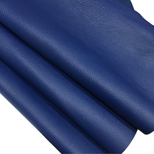 Pode incluir: Close-up de uma pilha de folhas de couro azul escuro. O couro tem uma superfície texturizada e é dobrado sobre si mesmo. A cor é um azul rico e saturado, e o material parece flexível e de alta qualidade.