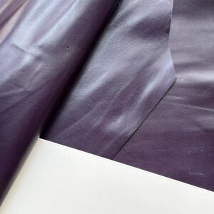 May include: A large, folded sheet of deep purple material with a smooth, semi-glossy finish and a subtle, fine-grained texture. The material is draped in soft, flowing folds, creating highlights and shadows across the dark violet surface.
