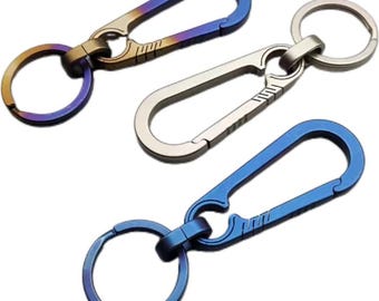 May include: Three titanium keychains with different color finishes. The keychains have a carabiner clip and a round ring. The colors are blue, gold, and a gradient of blue and gold.