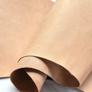Puede incluir: Primer plano de un rollo de cuero de color natural. El cuero es de color marrón claro con grano y textura visibles. Los bordes del cuero están ligeramente curvados, revelando la parte inferior rugosa.