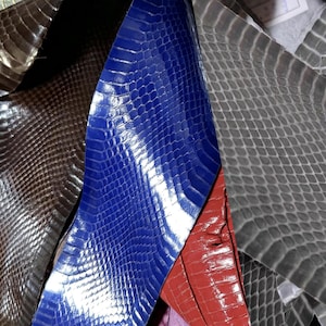 Könnte beinhalten: Eine Sammlung strukturierter Lederhäute in verschiedenen Farben. Das Bild zeigt ein leuchtendes Blau, ein tiefes Rot und ein metallisches Silberleder, alle mit einem schuppenartigen Muster. Weitere Häute in Grau- und Brauntönen sind ebenfalls sichtbar.