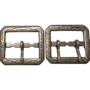 May include: Two antique silver-coloured belt buckles with intricate etched designs. Each buckle has a rectangular frame with a central bar and a prong for securing a belt. The buckles are displayed side-by-side, showcasing their detailed craftsmanship.