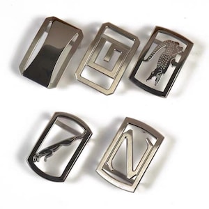 Può includere: Cinque fibbie per cintura argentate con diversi design. Le fibbie sono tutte rettangolari e hanno una finitura lucida. Una fibbia ha un semplice design geometrico, un'altra ha un coccodrilo, un'altra ha un giaguaro, un'altra ha una lettera N e l'ultima ha una superficie liscia.