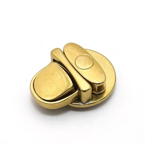 May include: A gold-toned metal clasp with a rounded, oval-shaped design. The clasp has a hinged mechanism and a small, circular button.