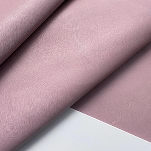 Puede incluir: Primer plano de una hoja doblada de cuero sintético de color malva. El material tiene una veta sutil y texturizada y un acabado mate. La tela está drapeada sobre una superficie blanca, creando sombras y resaltando la textura.