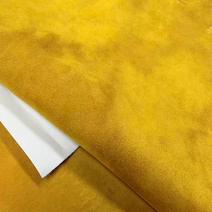 May include: A mustard yellow fabric with a soft, suede-like texture. The material is folded over to reveal a smooth, white underside with a serrated, zigzag-cut edge. The surface of the yellow fabric shows subtle variations in tone and light.