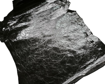 Puede incluir: Una hoja cuadrada y delgada de metal plateado con una textura superficial arrugada y altamente reflectante. El material parece delicado y ligero, presentando numerosos pliegues y arrugas finas que captan la luz sobre su acabado oscuro y brillante.
