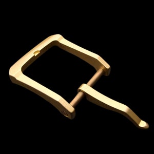 May include: A gold-toned metal buckle with a rectangular frame and a curved, hinged clasp. The buckle is designed for a strap or belt.